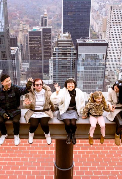 Visitors sitting on the edge at Top of the Rock, New York City skyline in the background.