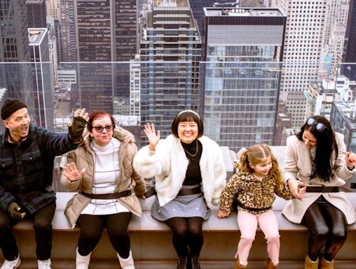 Visitors sitting on the edge at Top of the Rock, New York City skyline in the background.