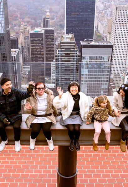 Visitors sitting on the edge at Top of the Rock, New York City skyline in the background.
