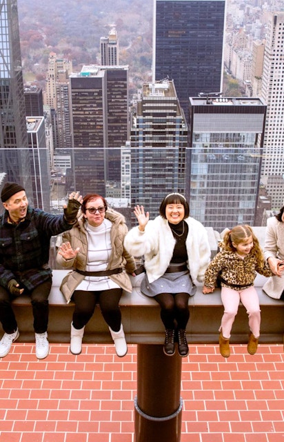 Visitors sitting on the edge at Top of the Rock, New York City skyline in the background.