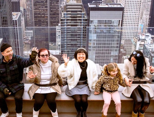 Visitors sitting on the edge at Top of the Rock, New York City skyline in the background.