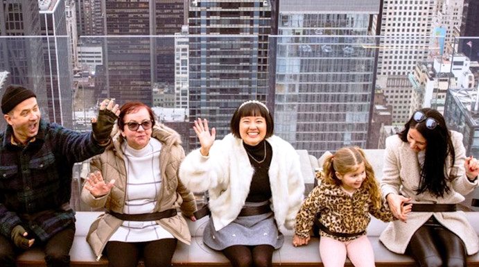 Visitors sitting on the edge at Top of the Rock, New York City skyline in the background.