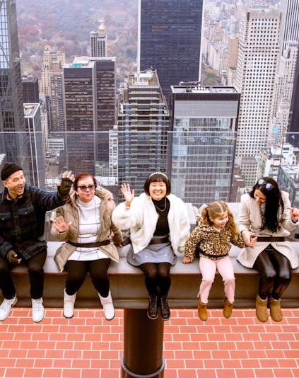 Visitors sitting on the edge at Top of the Rock, New York City skyline in the background.