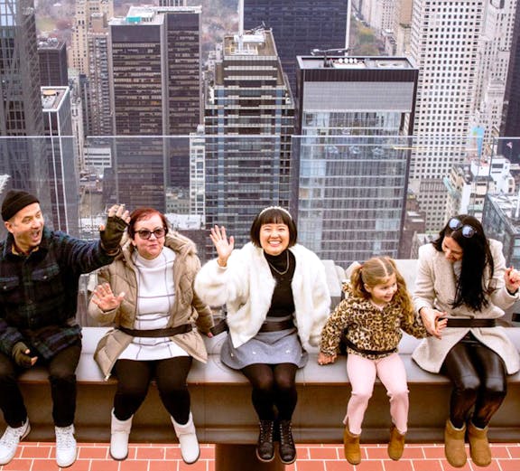 Visitors sitting on the edge at Top of the Rock, New York City skyline in the background.