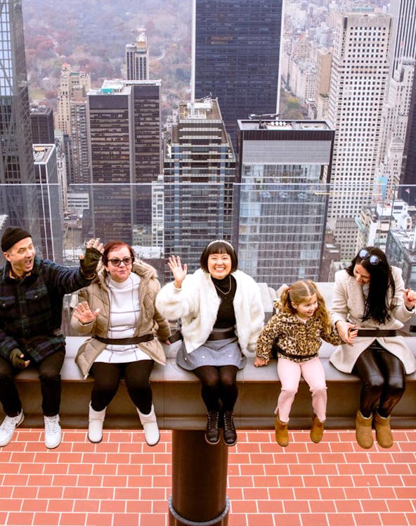 Visitors sitting on the edge at Top of the Rock, New York City skyline in the background.