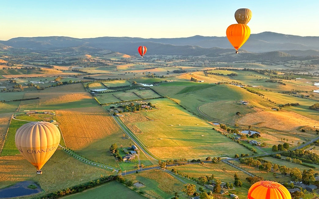 Hot air balloons floating over Yarra Valley at sunrise, showcasing expansive vineyards and rolling hills.