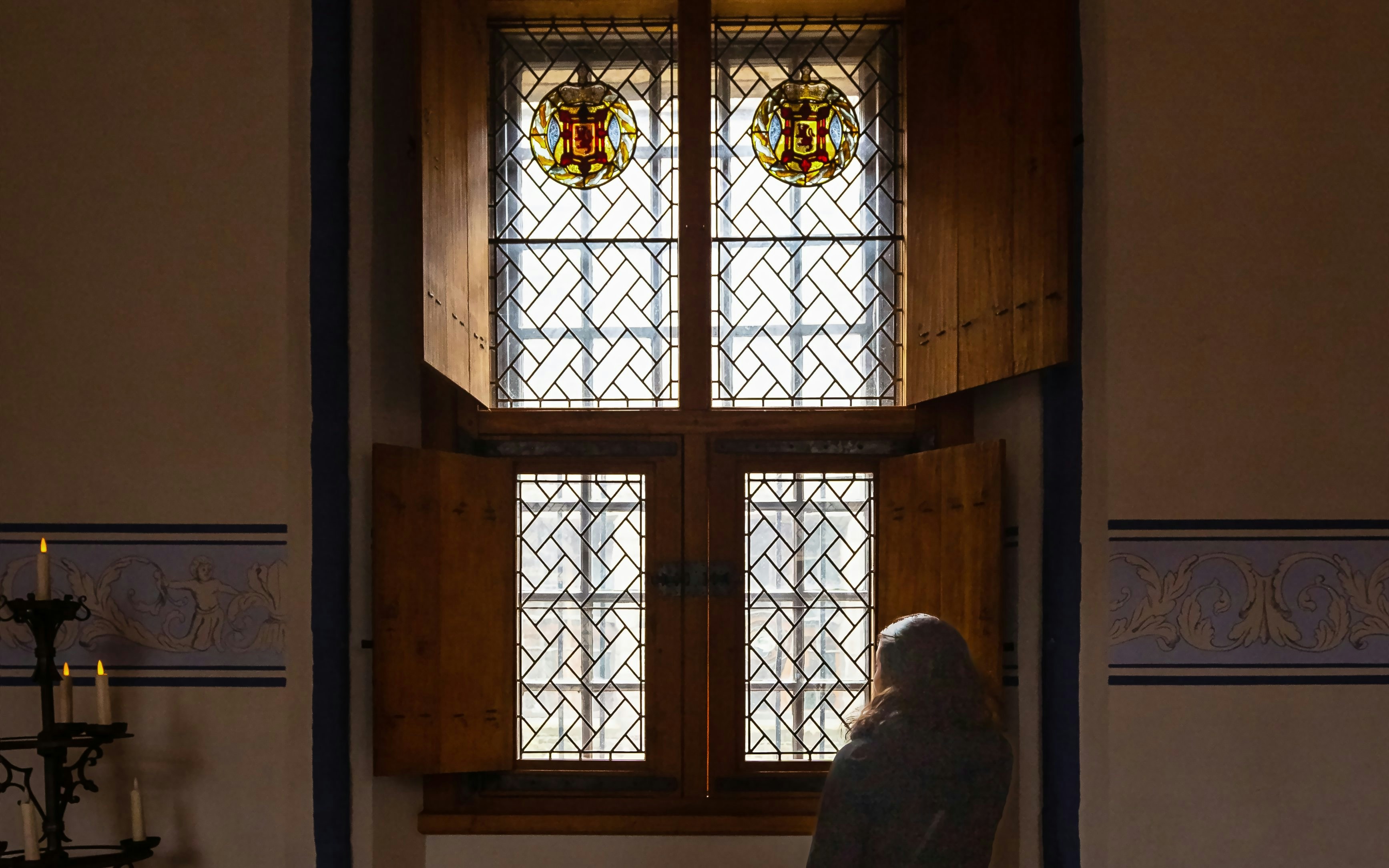 Person gazing out of a stained glass castle window with wooden shutters.