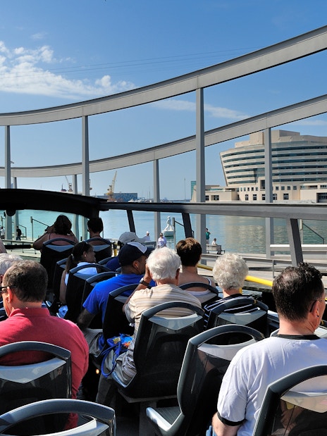 Passengers on Barcelona City Hop On Hop Off Tour bus near waterfront.