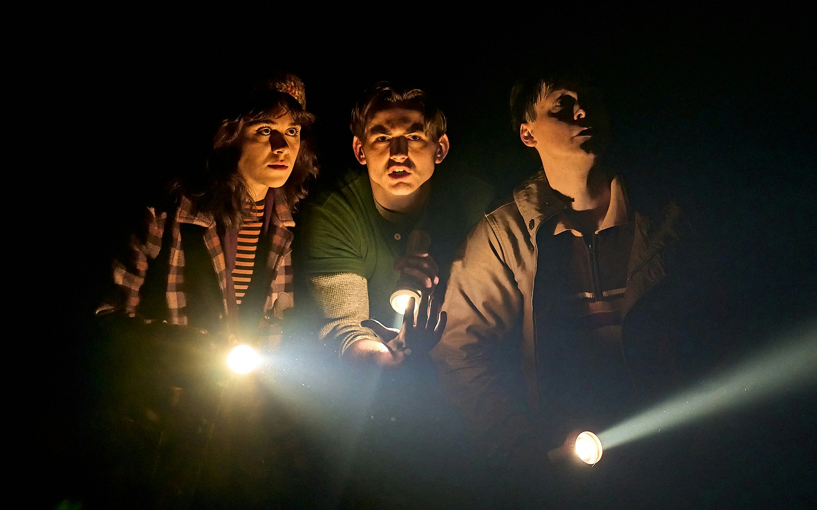 Three people holding flashlights in a dark setting, related to Stranger Things: The First Shadow.