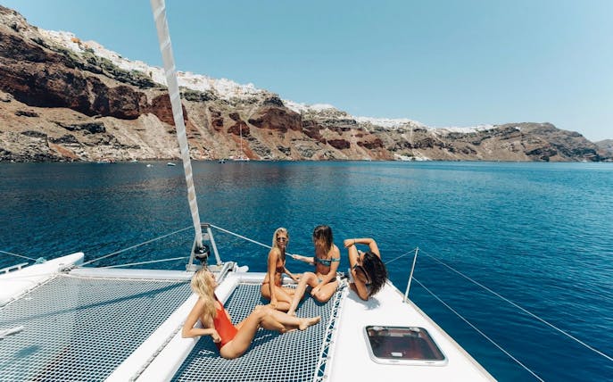 Catamaran cruise in Santorini's caldera with people relaxing on deck, cliffs in background.
