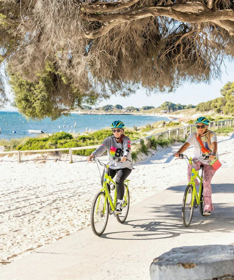 Cyclists on a path by the beach on Rottnest Island, with ocean view and boats in the background.