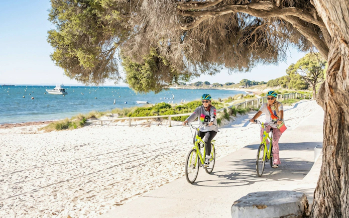 Cyclists on a path by the beach on Rottnest Island, with ocean view and boats in the background.