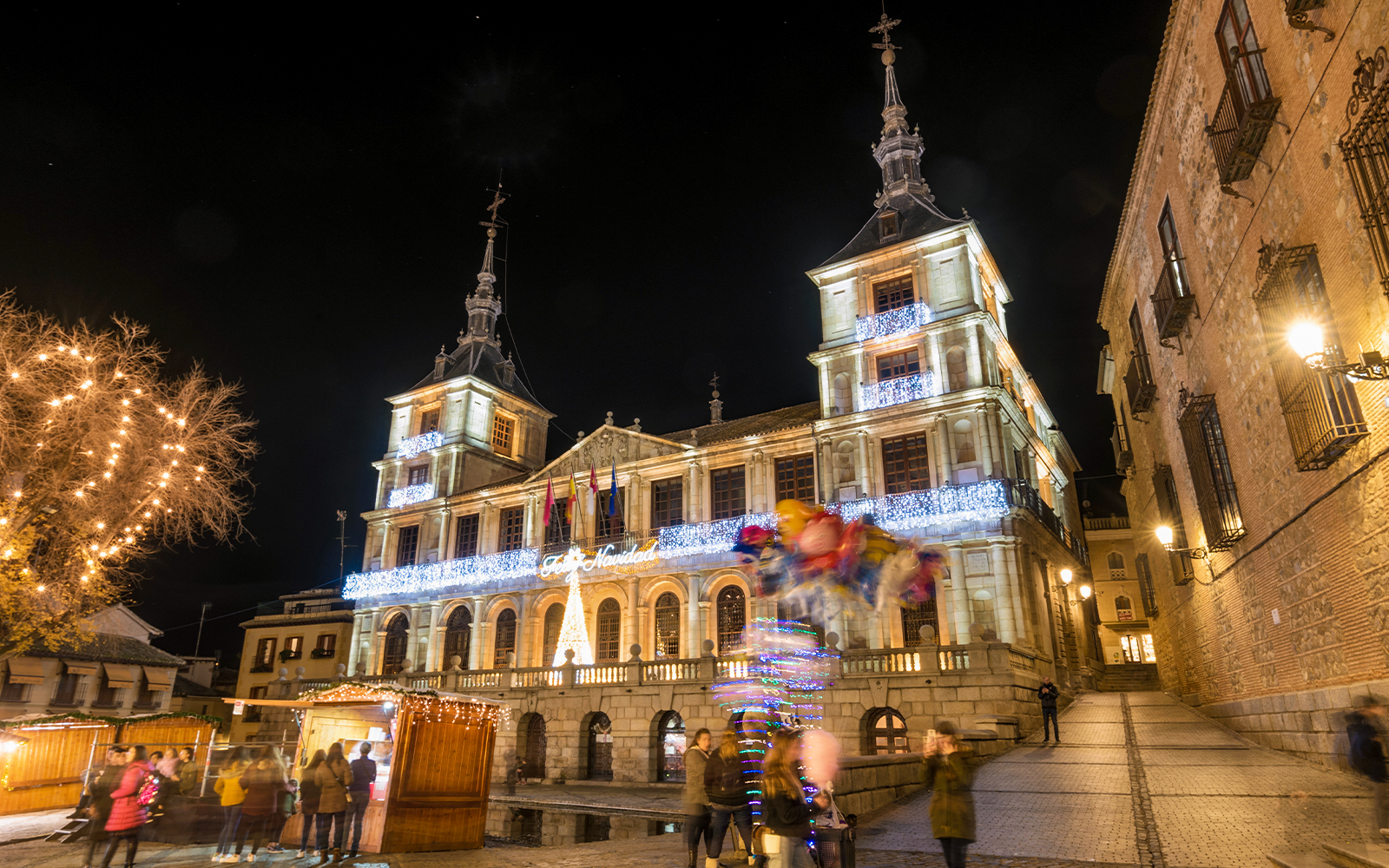 Toledo town hall illuminated with Christmas lights at night, festive market stalls below.