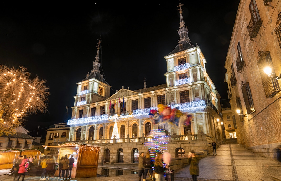 Toledo town hall illuminated with Christmas lights at night, festive market stalls below.