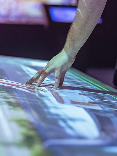 Interactive display showing Montserrat Monastery tour details with hand pointing.