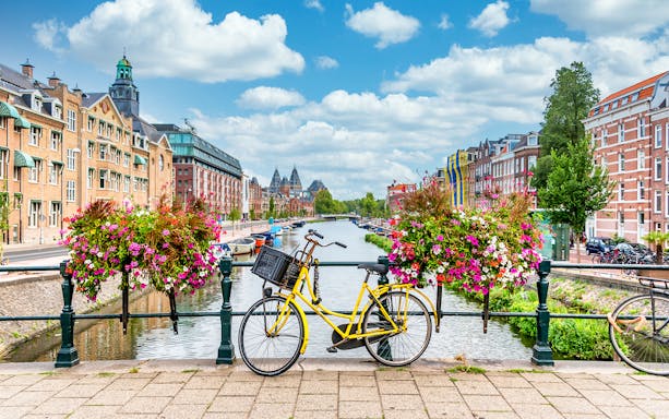 Yellow bicycle on a bridge over a canal in Amsterdam with flowers and historic buildings.