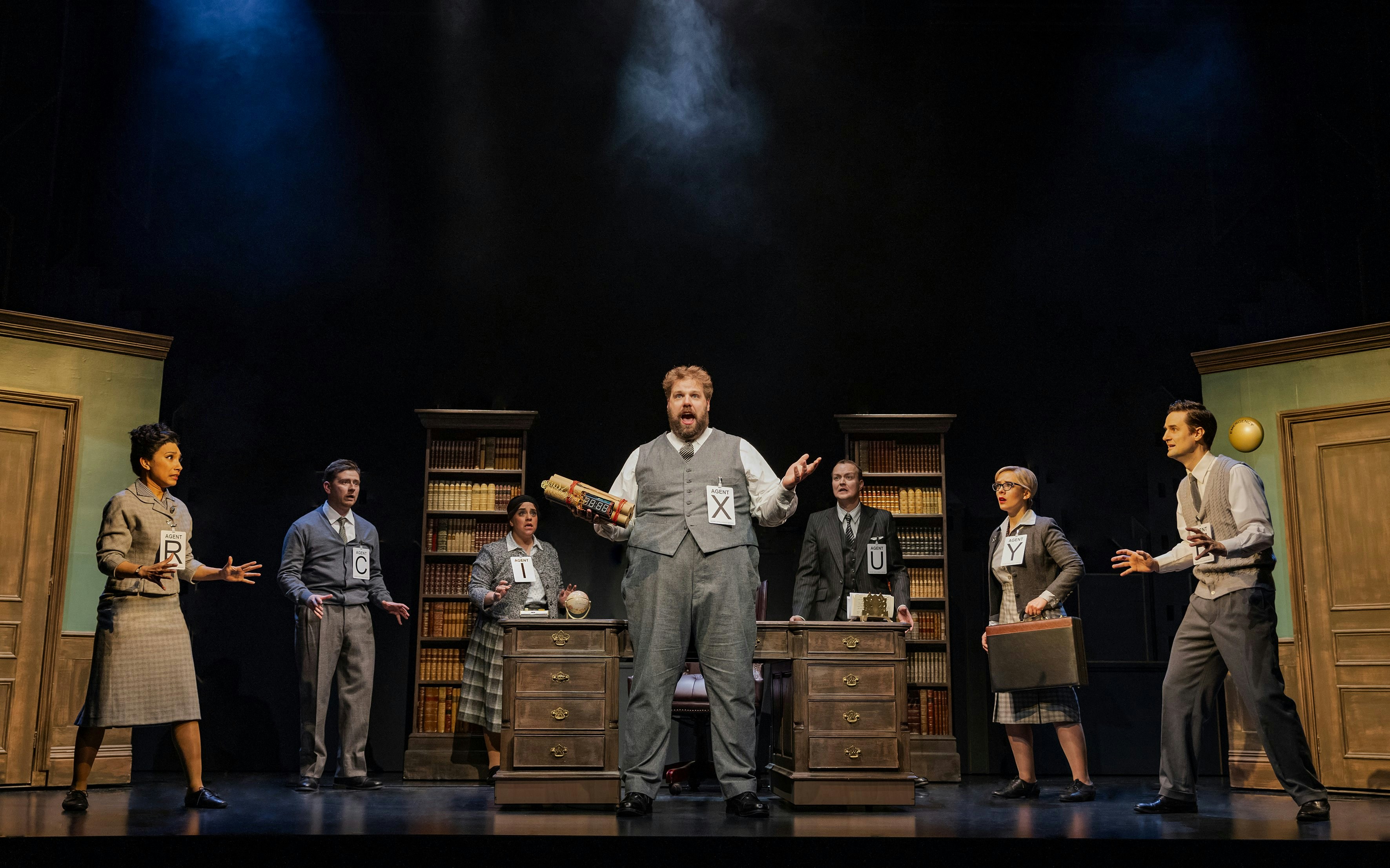 Actors on stage in "The Comedy About Spies" with spy-themed props and costumes.