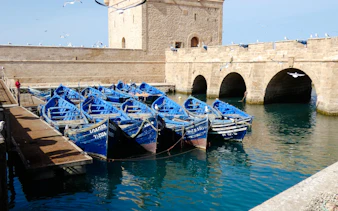 Blue fishing boats docked at Essaouira harbor with stone fortress in the background.