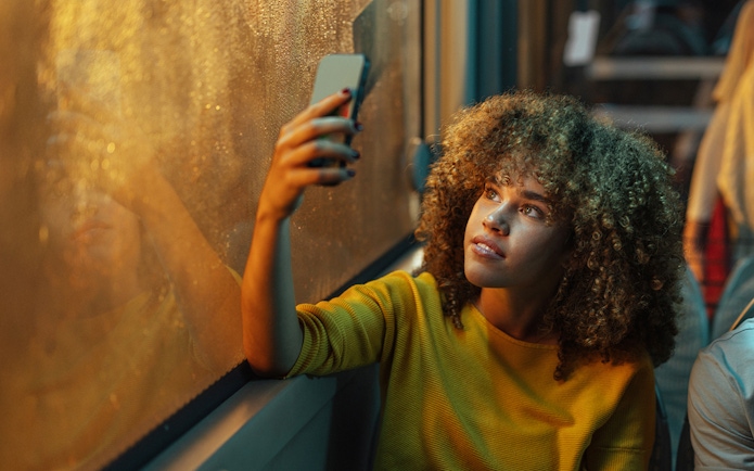 Young woman looking out bus window at night, holding a smartphone.