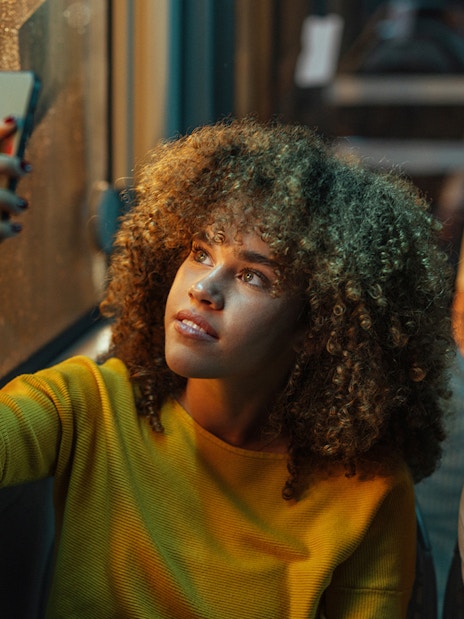 Young woman looking out bus window at night, holding a smartphone.