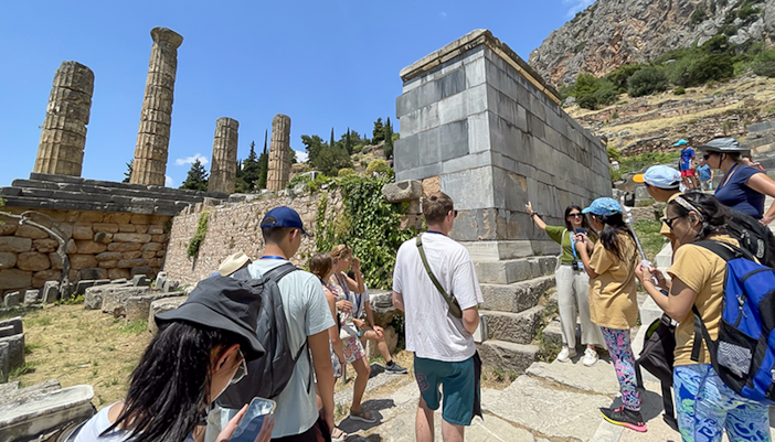 Tourists listening to guide at ancient ruins in Delphi, Greece during day trip.