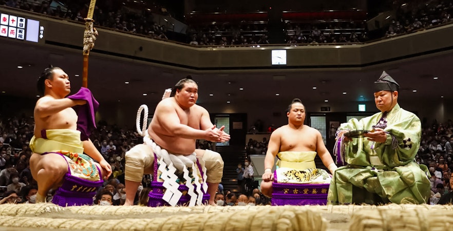 Sumo wrestlers and referee in traditional attire at Tokyo Grand Sumo Tournament.