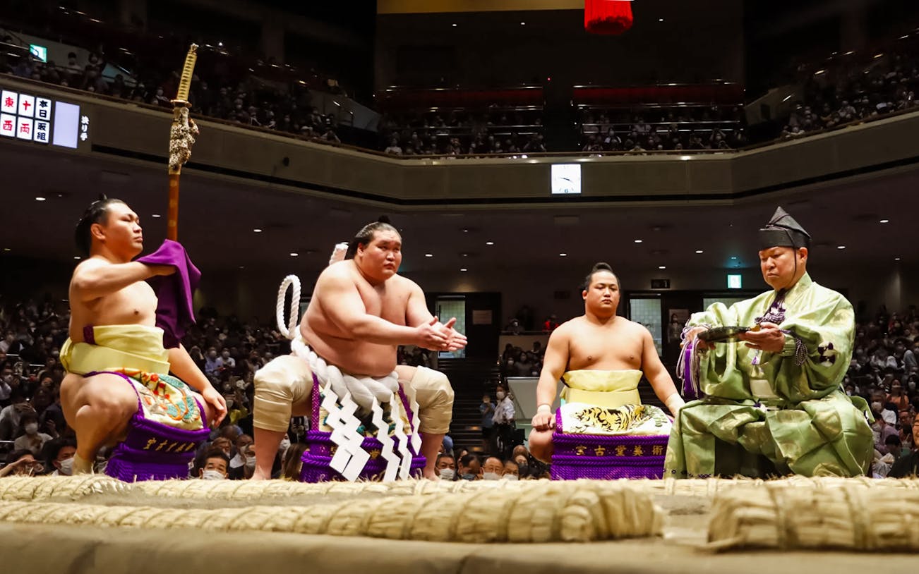 Sumo wrestlers and referee in traditional attire at Tokyo Grand Sumo Tournament.