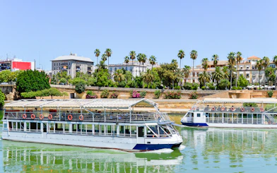 Sevilla city cruise boats on the Guadalquivir River with palm trees and buildings in the background.