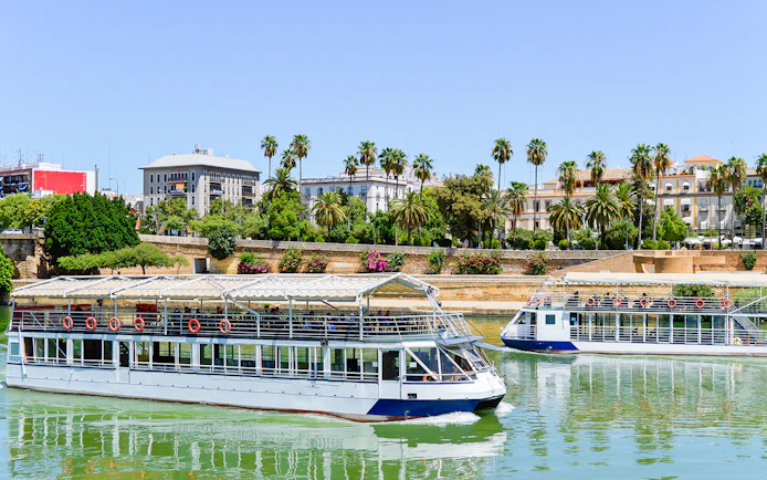 Sevilla city cruise boats on the Guadalquivir River with palm trees and buildings in the background.