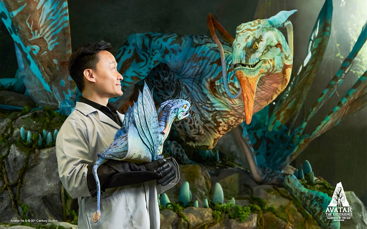 Man holding a small dragon-like creature with a larger one in the background at Gardens by the Bay, Singapore.