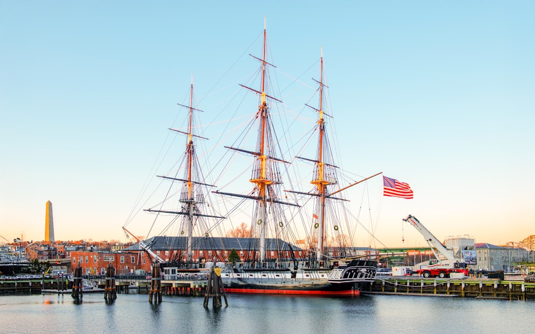 USS Constitution docked in Boston with American flag and cityscape in the background.