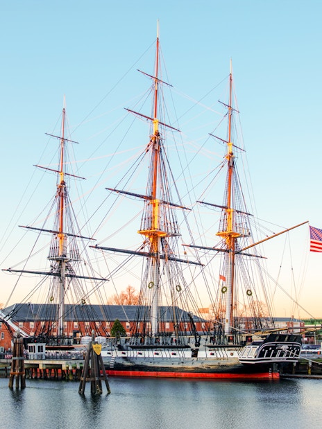 USS Constitution docked in Boston with American flag and cityscape in the background.