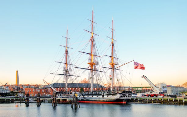 USS Constitution docked in Boston with American flag and cityscape in the background.