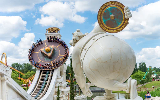 Visitors on a spinning ride at Parc Astérix, Paris, with a large statue in the foreground.