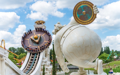 Visitors on a spinning ride at Parc Astérix, Paris, with a large statue in the foreground.