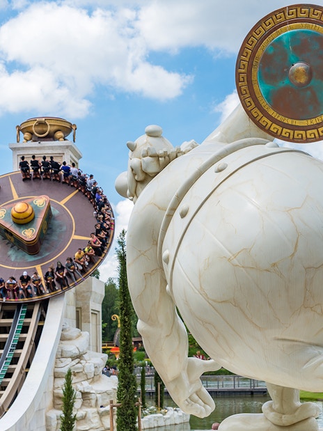 Visitors on a spinning ride at Parc Astérix, Paris, with a large statue in the foreground.