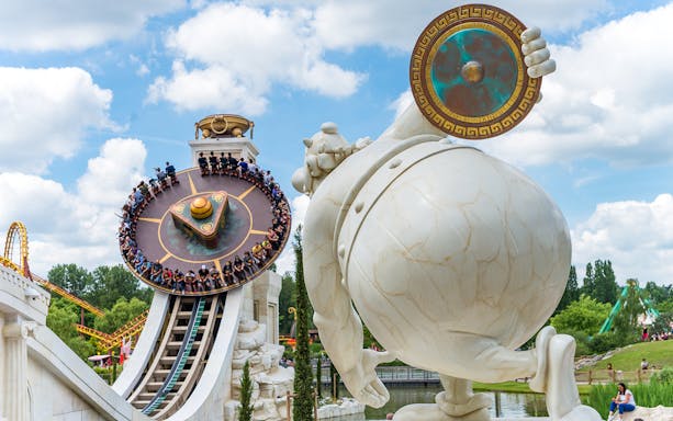 Visitors on a spinning ride at Parc Astérix, Paris, with a large statue in the foreground.