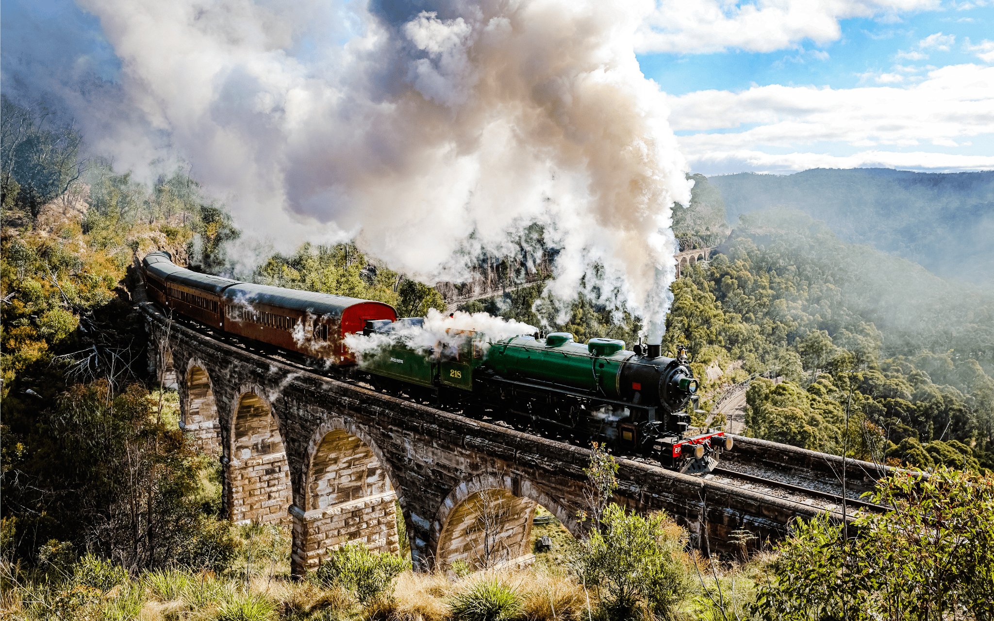 Steam train crossing viaduct in Blue Mountains, Australia, surrounded by lush forest.