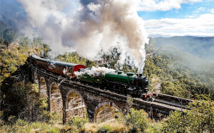 Steam train crossing viaduct in Blue Mountains, Australia, surrounded by lush forest.