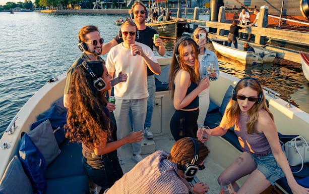 People enjoying a party cruise on a canal in Amsterdam, wearing headphones and holding drinks.
