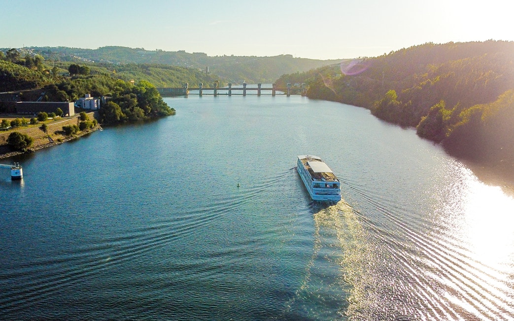Aerial view of a boat cruising on the Douro River with hills and a bridge in the background.