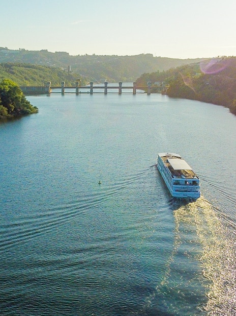 Aerial view of a boat cruising on the Douro River with hills and a bridge in the background.