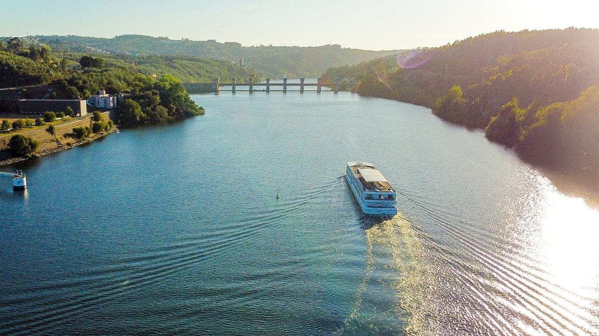 Aerial view of a boat cruising on the Douro River with hills and a bridge in the background.