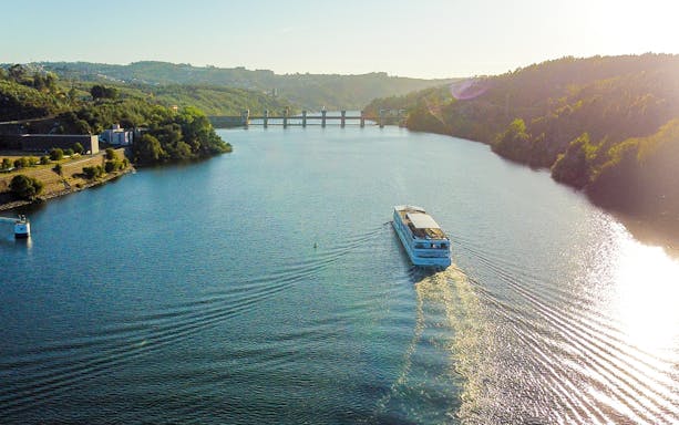 Aerial view of a boat cruising on the Douro River with hills and a bridge in the background.