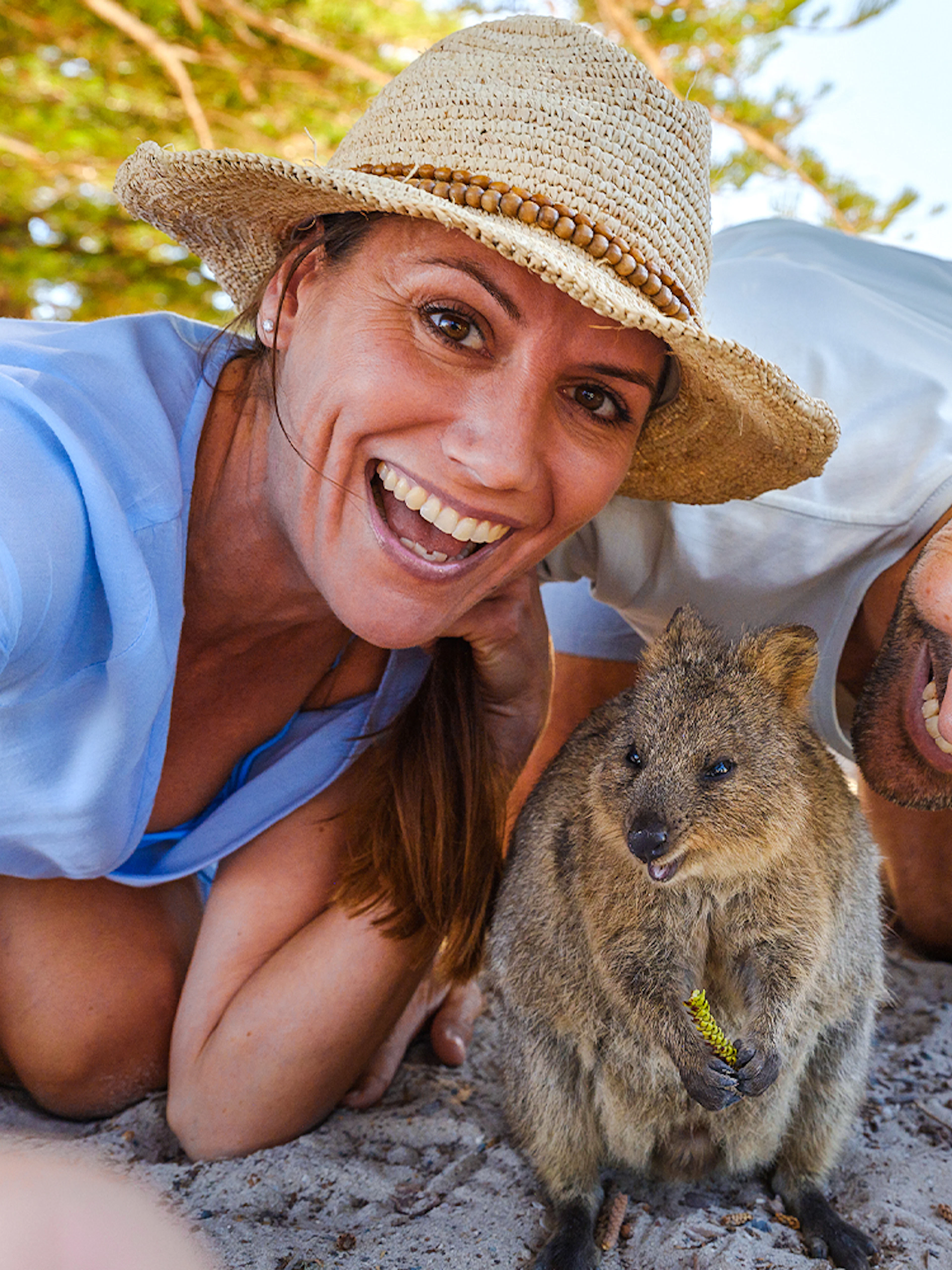Tourists smiling with a quokka on Rottnest Island, bicycles and beach in background.