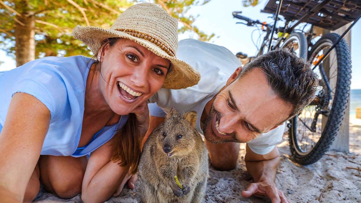 Tourists smiling with a quokka on Rottnest Island, bicycles and beach in background.