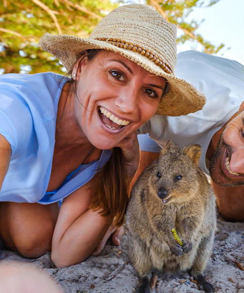 Tourists smiling with a quokka on Rottnest Island, bicycles and beach in background.