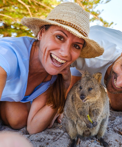Tourists smiling with a quokka on Rottnest Island, bicycles and beach in background.