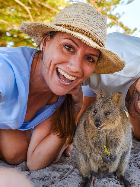 Tourists smiling with a quokka on Rottnest Island, bicycles and beach in background.