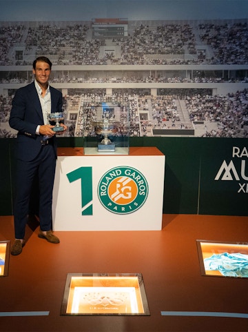 Man holding tennis trophies at Rafa Nadal Museum, Mallorca, with Roland Garros display in background.